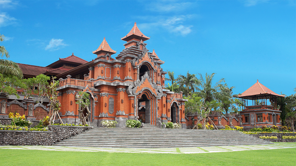 Front view of a red-brick temple with ornate arches, stair steps, and spired towers under a bright blue sky.