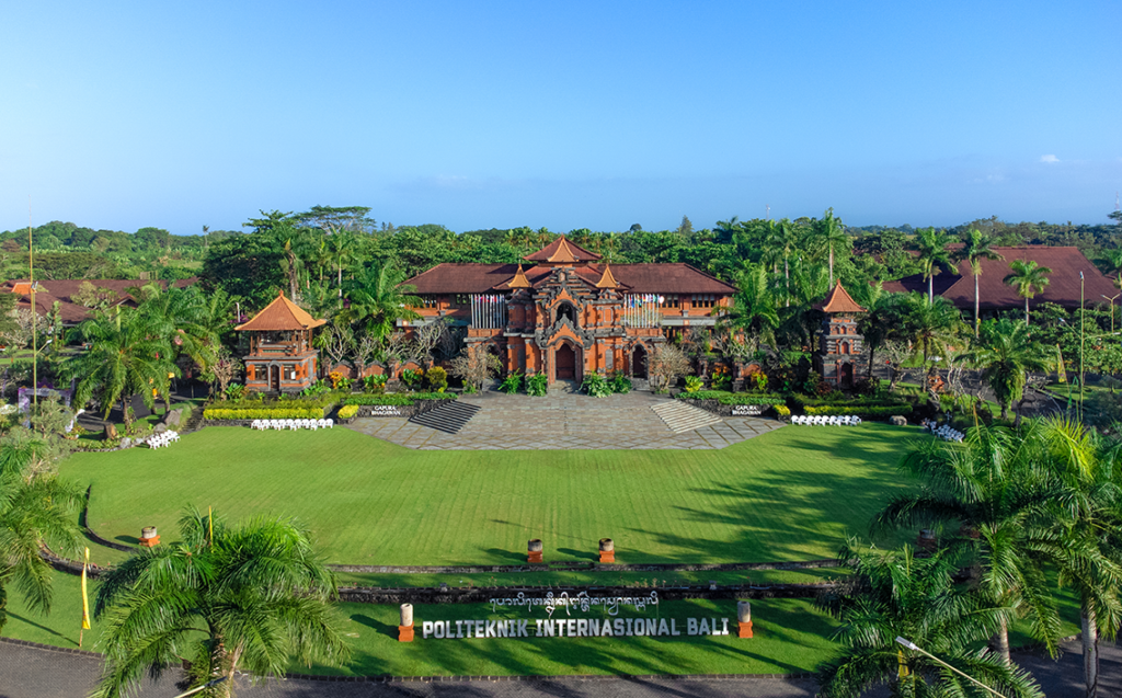 Aerial view of a tropical college campus with red brick buildings, green lawn, and palm trees; sign reads POLITEKNIK INTERNASIONAL BALI at the entrance