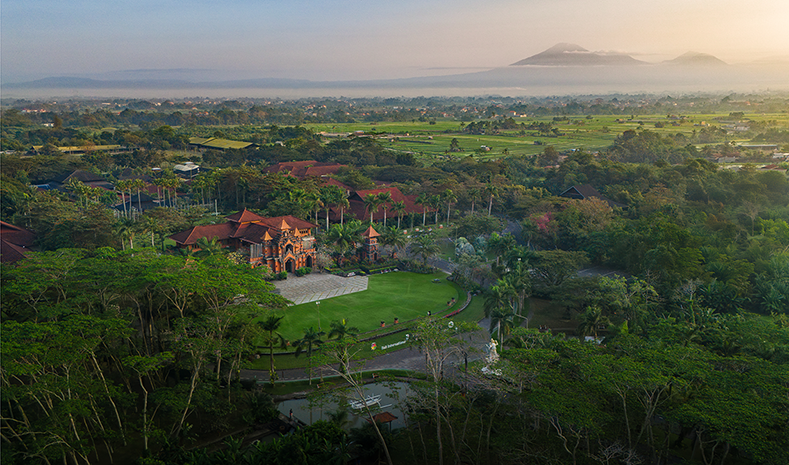 Aerial view of a tropical resort with red-roof buildings, lush greenery, and distant mountains at sunrise.